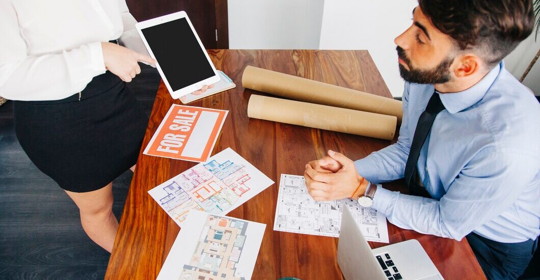 Real estate agent presenting property details to a potential buyer with floor plans, a 'For Sale' sign, and a digital tablet in an office setting.