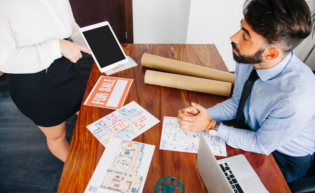 Real estate agent presenting property details to a potential buyer with floor plans, a 'For Sale' sign, and a digital tablet in an office setting.
