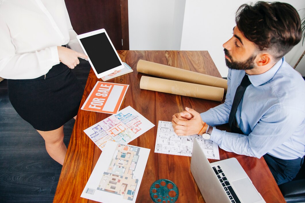 Real estate agent presenting property details to a potential buyer with floor plans, a 'For Sale' sign, and a digital tablet in an office setting.
